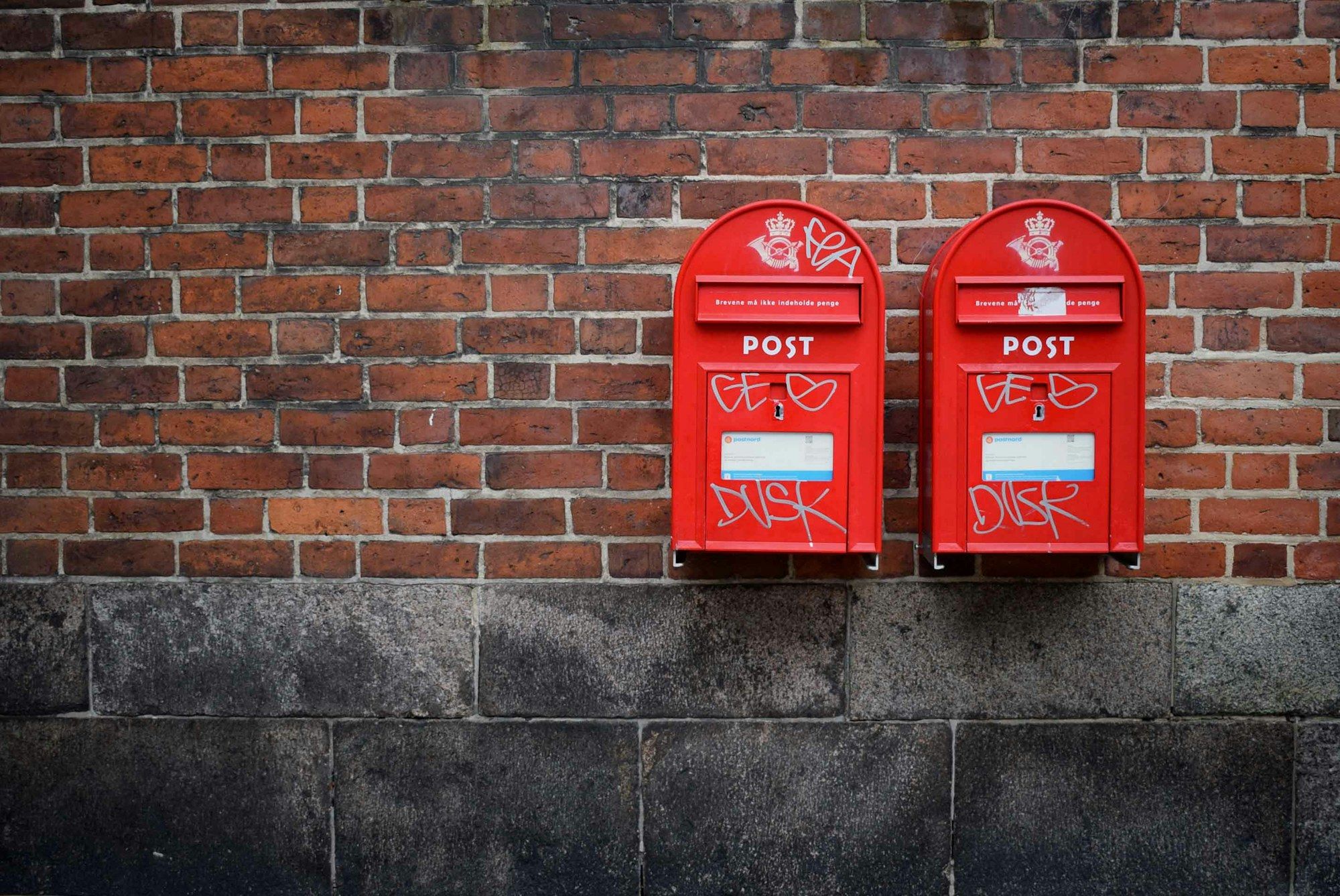 Dos buzones de correo rojos montados en un muro de ladrillo, cerrados, con la ranura de entrada a la vista y ninguna señal de lo que hay dentro.